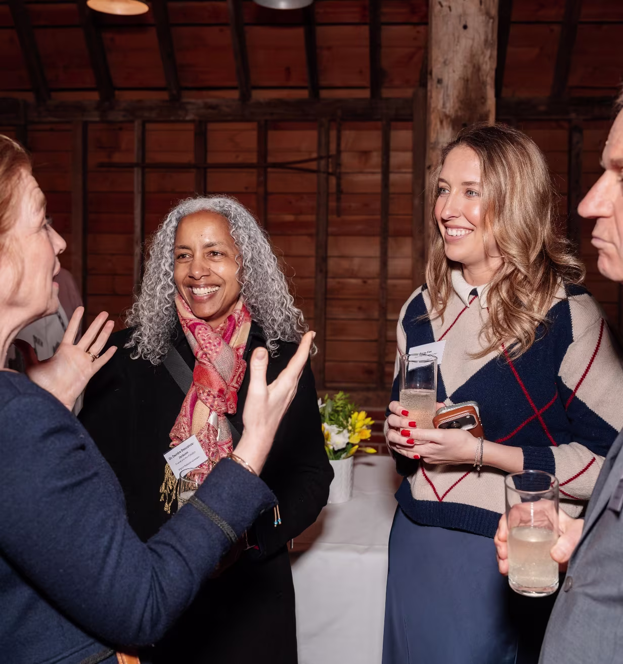 Four people smiling and talking indoors at a social event, holding drinks with a wooden wall background.