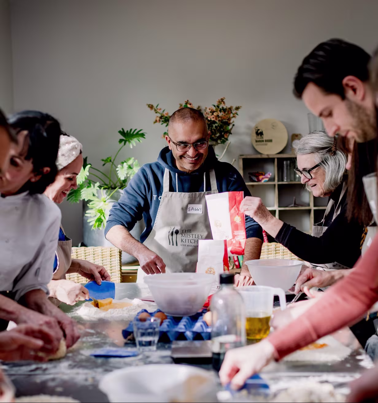 Group of people happily baking together around a table with flour, eggs, and ingredients.