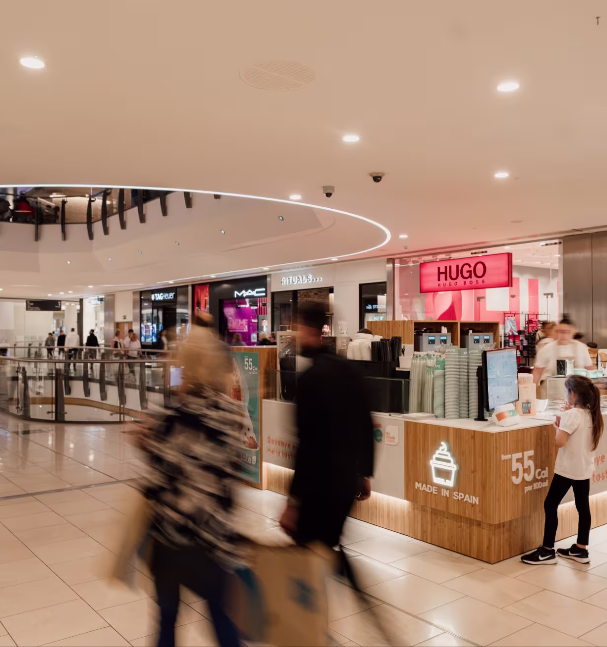Busy shopping mall corridor with blurred shoppers and a Made in Spain kiosk in front of Hugo Boss store.