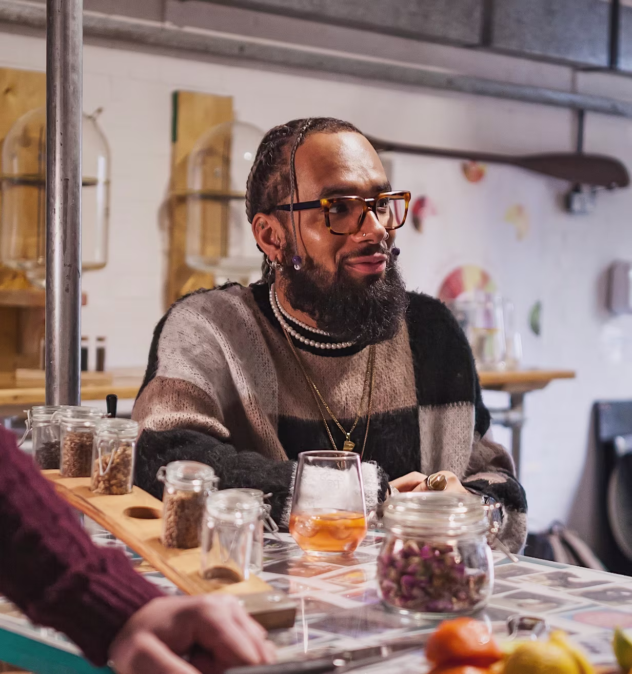 Man with glasses and beard sitting at a table with jars of spices and a drink in a cozy setting.