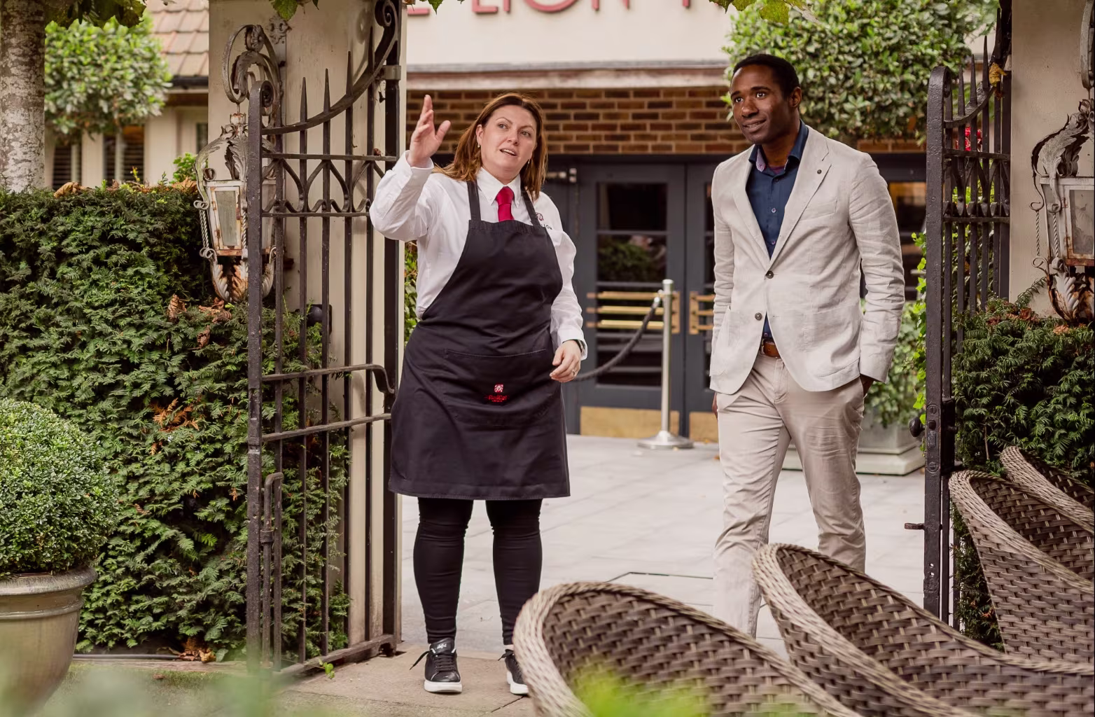 Woman in black apron gesturing while talking to man in beige suit near restaurant entrance.