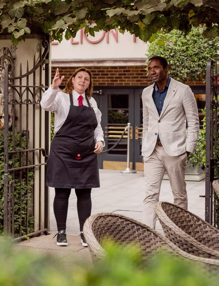 Woman in apron gestures as man in light suit listens in an outdoor seating area at a restaurant.