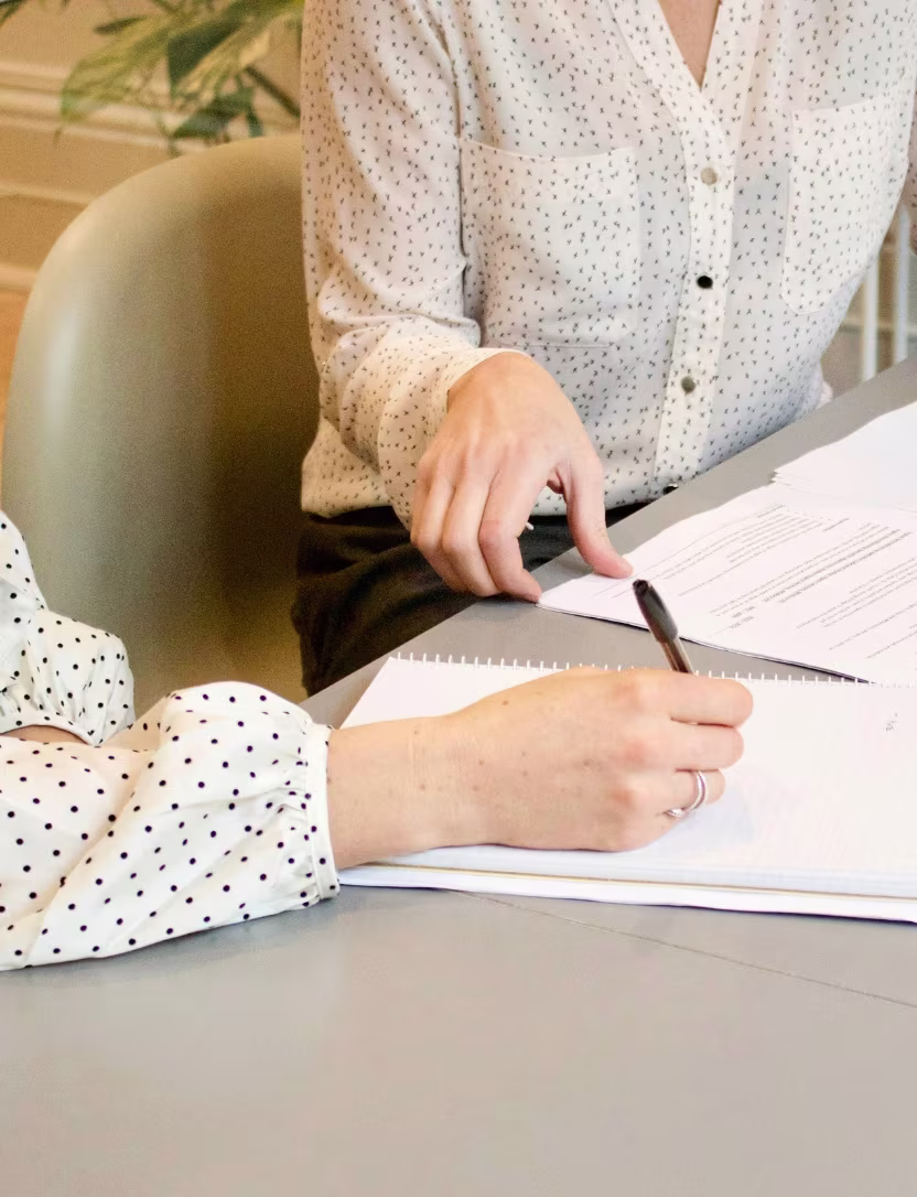 Person in polka dot sleeve writing on notepad while another person points at documents on a table.
