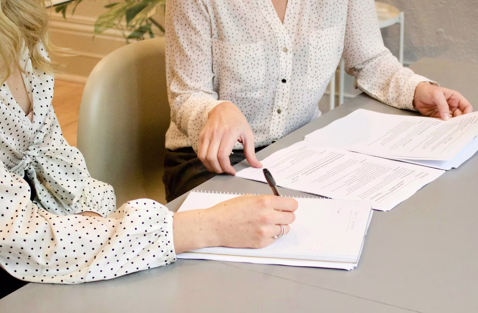 Two people in white polka-dot shirts reviewing and writing on business documents at a grey table.