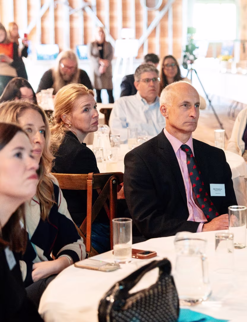 Group of adults listening attentively at a formal indoor event with tables and drinks.