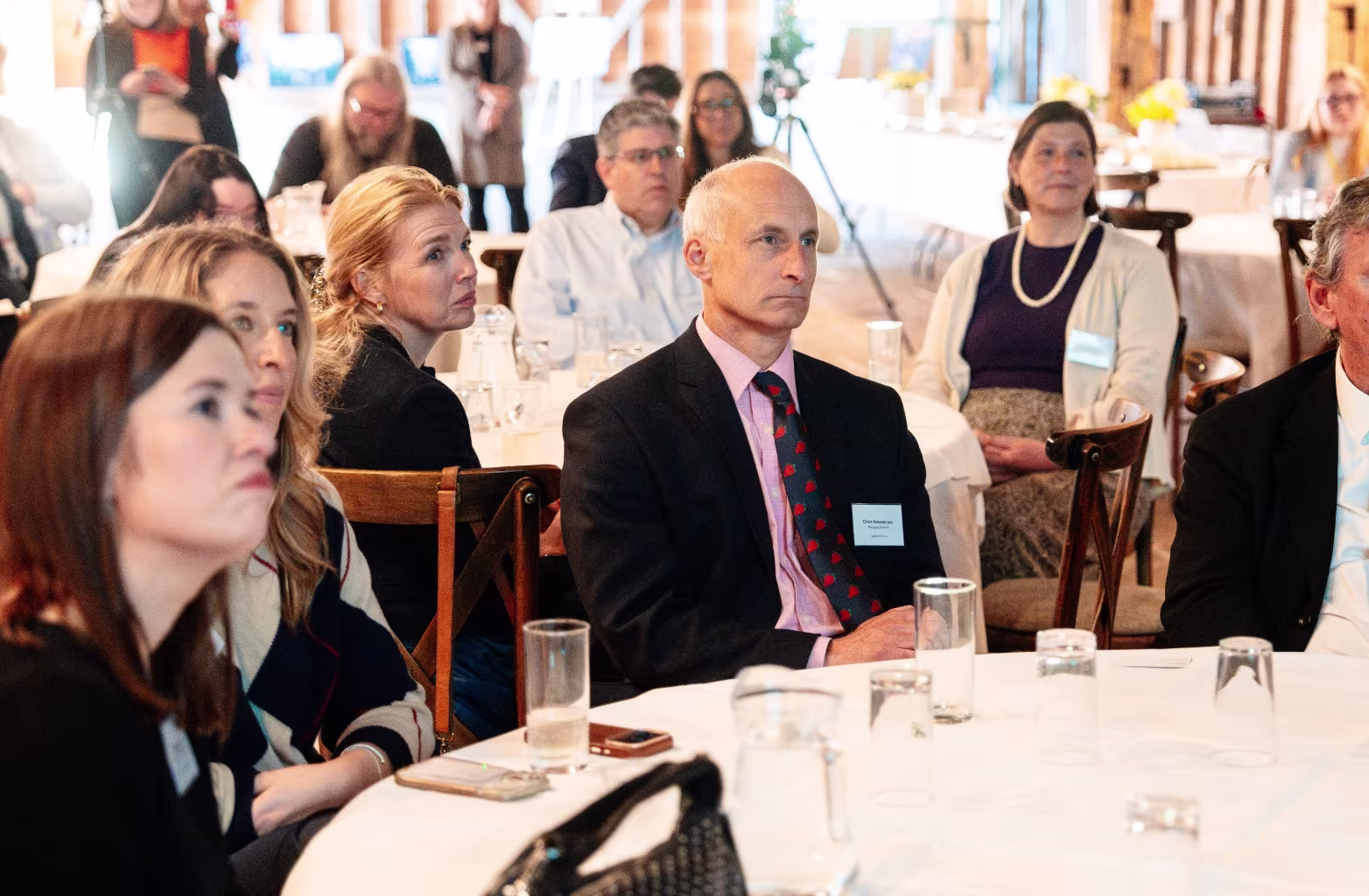 Diverse group of adults attentively listening at a formal event sitting around tables with glasses.