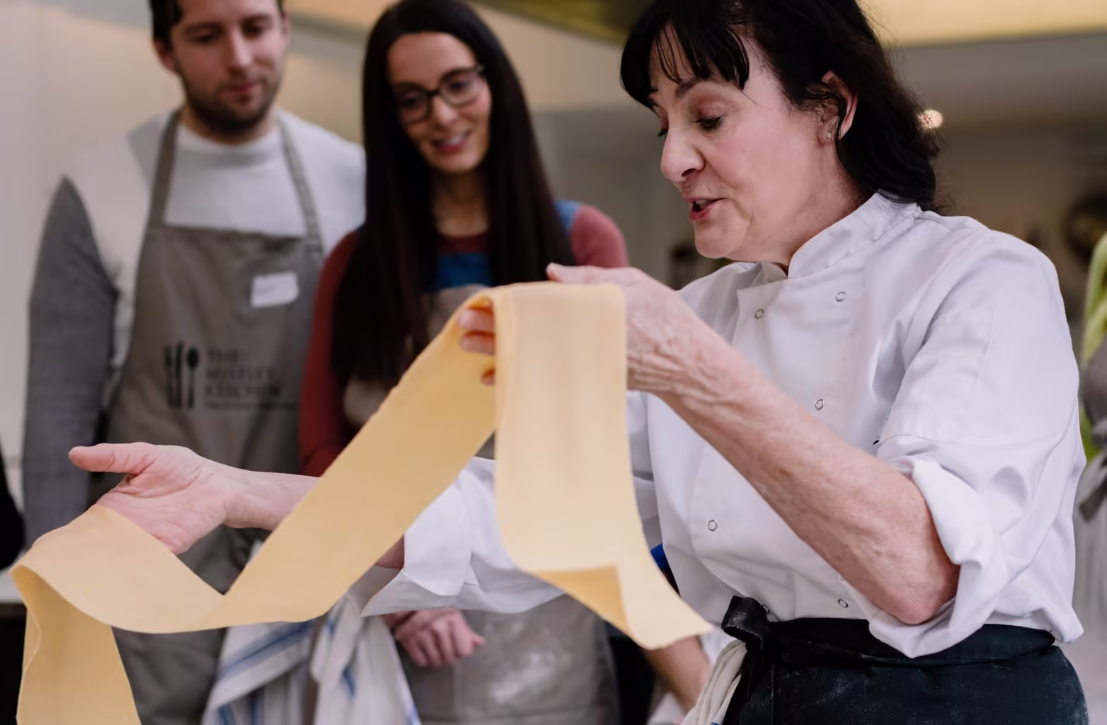 Woman chef holding and showing a long sheet of fresh pasta to two people watching attentively.