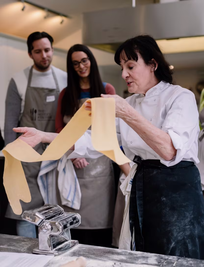 Woman chef demonstrating how to roll out fresh pasta dough with a pasta machine while two people watch.