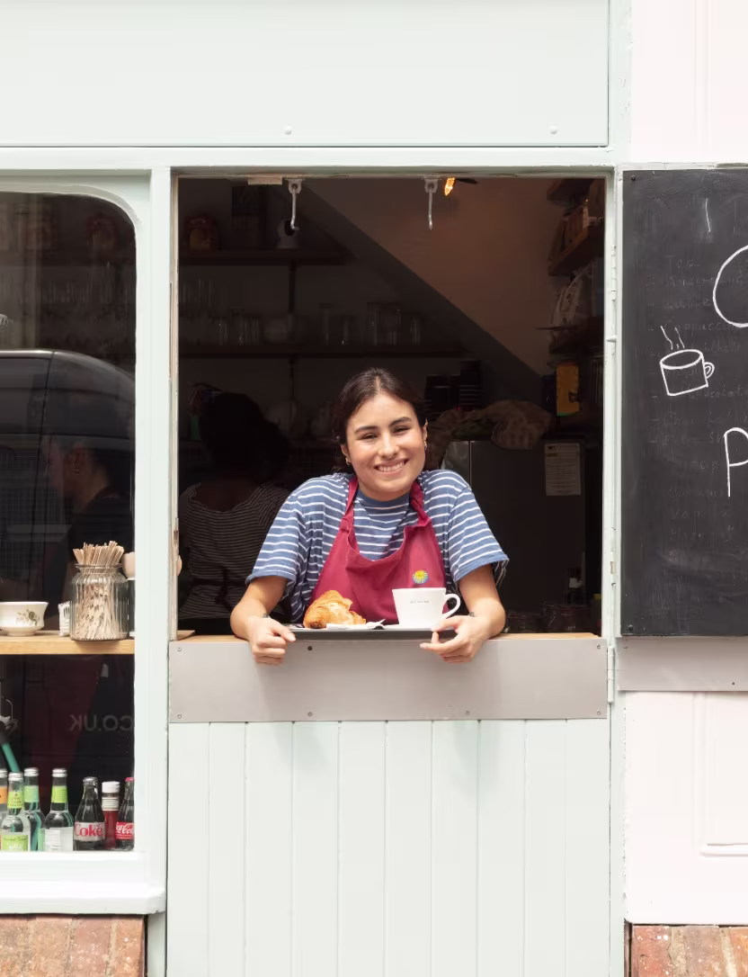 Smiling woman in apron serving coffee and croissant from cafe window.