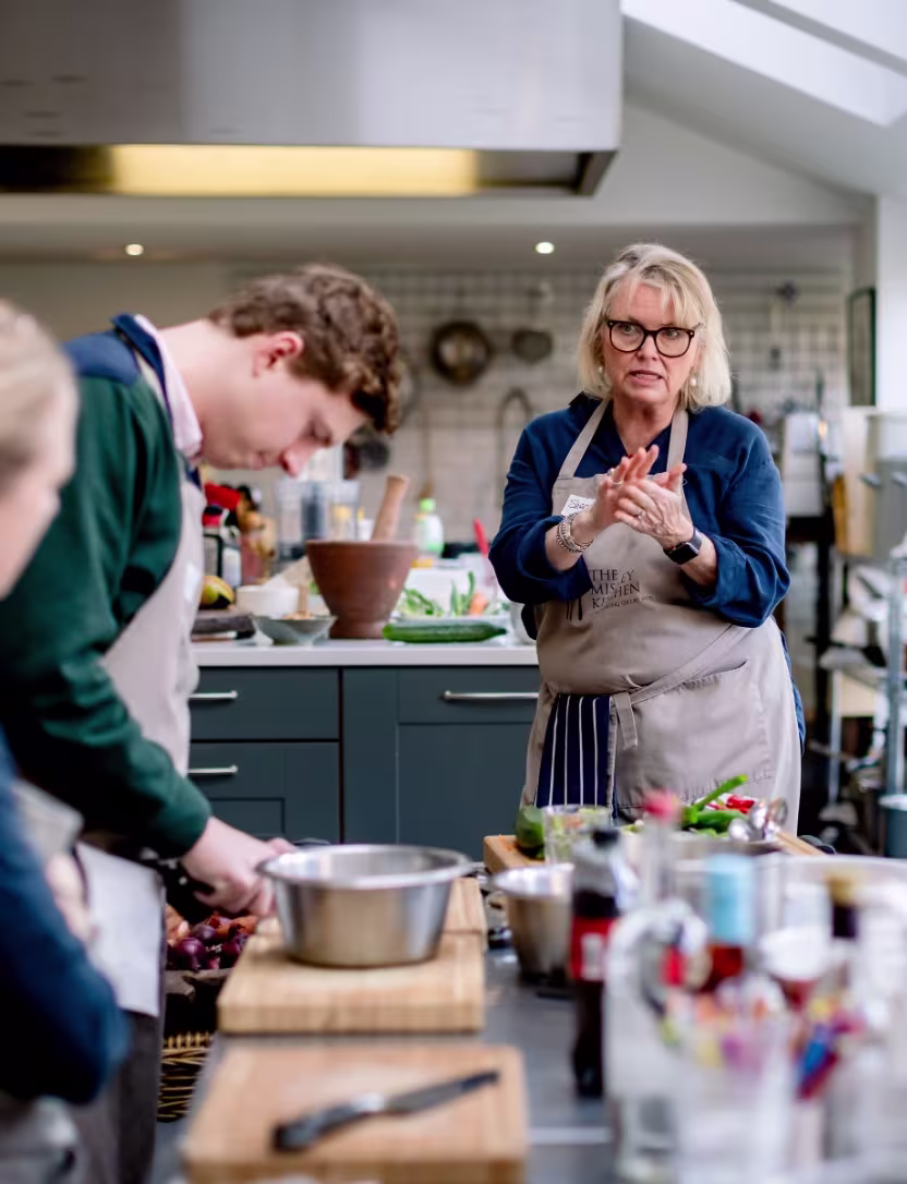 Woman instructing in a kitchen class while a man chops food at a counter.