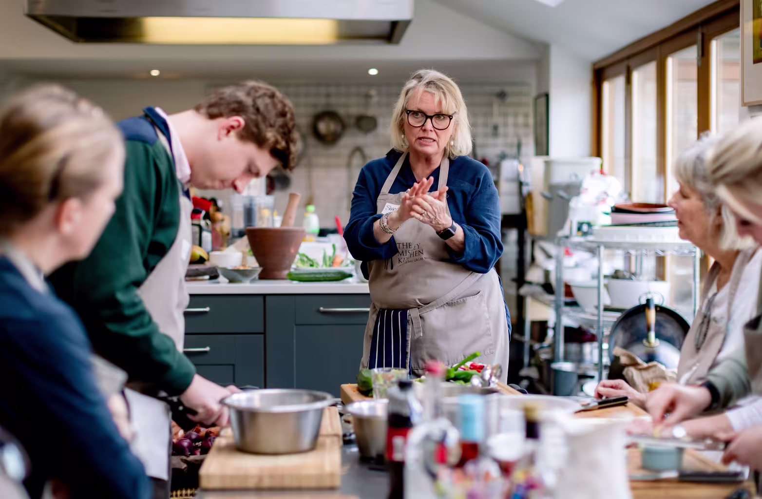 Woman chef instructs group in kitchen, with others preparing food on counters around her.