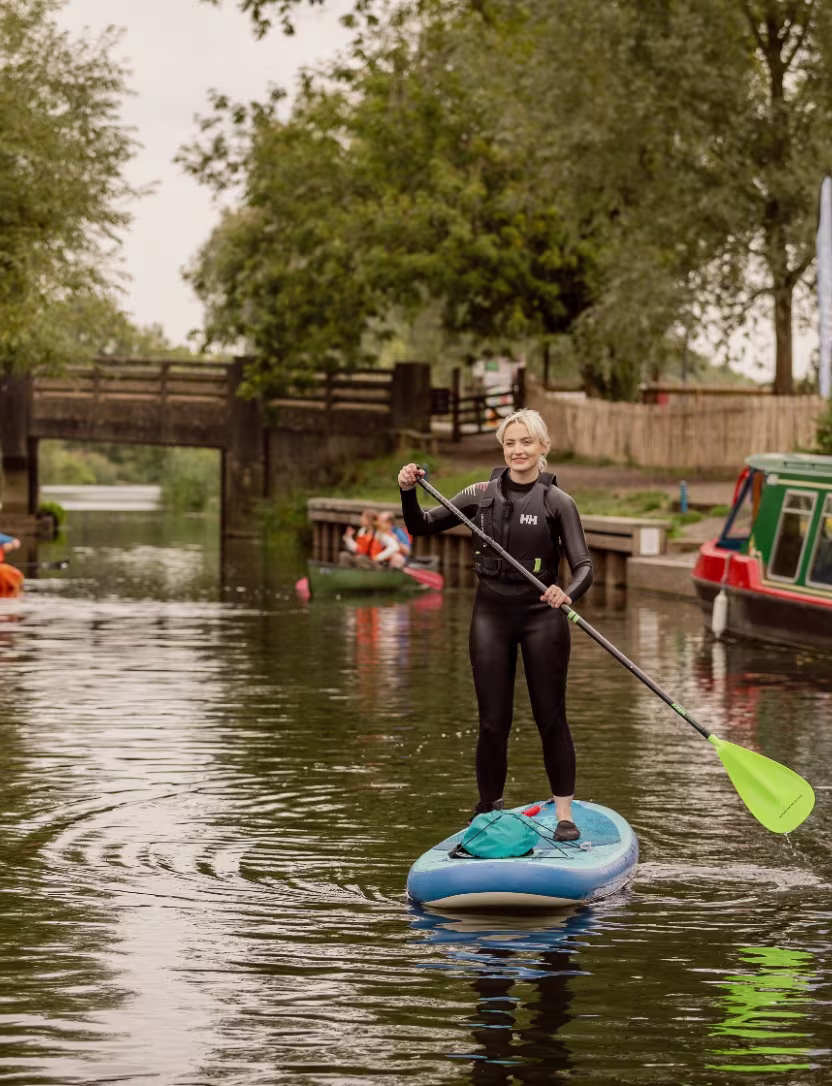 Woman wearing wetsuit paddleboarding on a calm canal with boats and trees in the background.