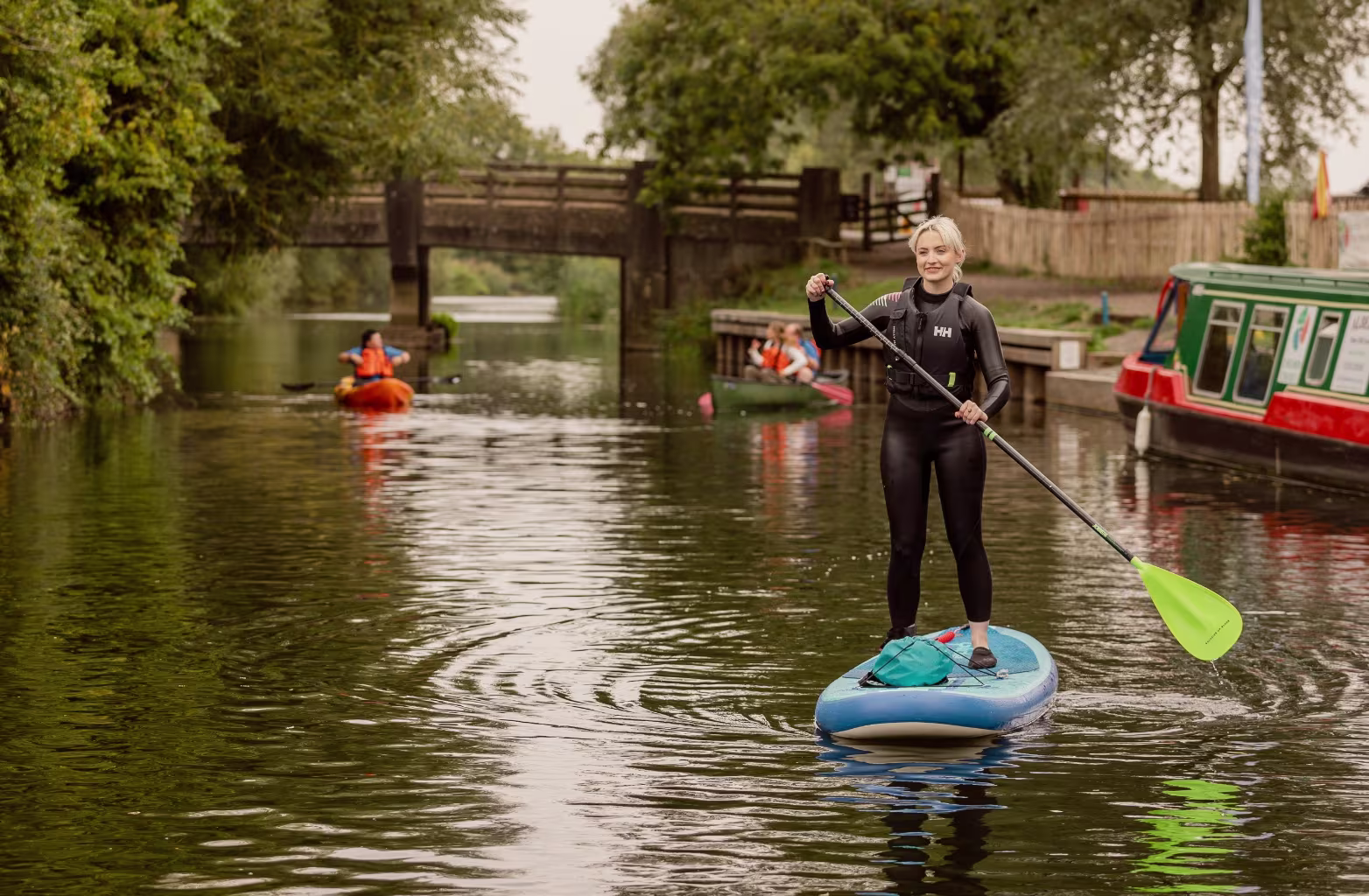 Woman in wetsuit paddleboarding on a calm canal with kayakers and a red green boat nearby.