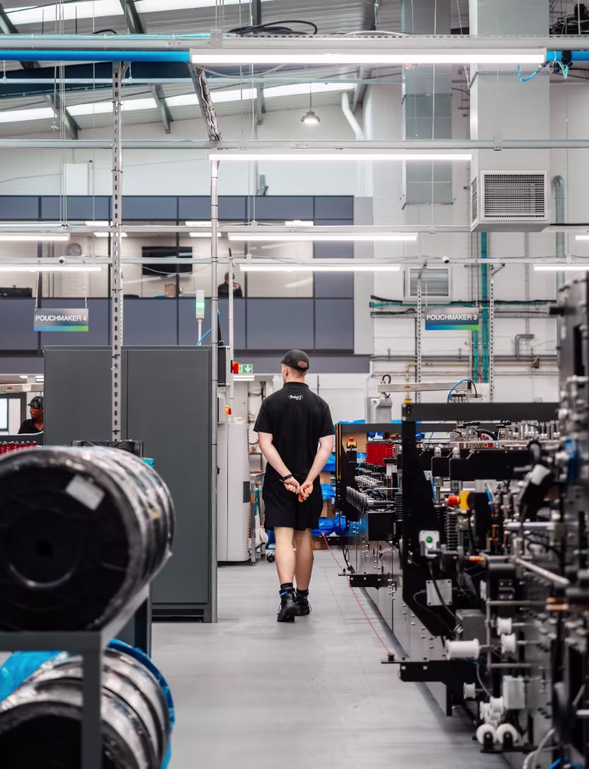 Factory worker in black uniform walking down aisle between machines and equipment in industrial setting.
