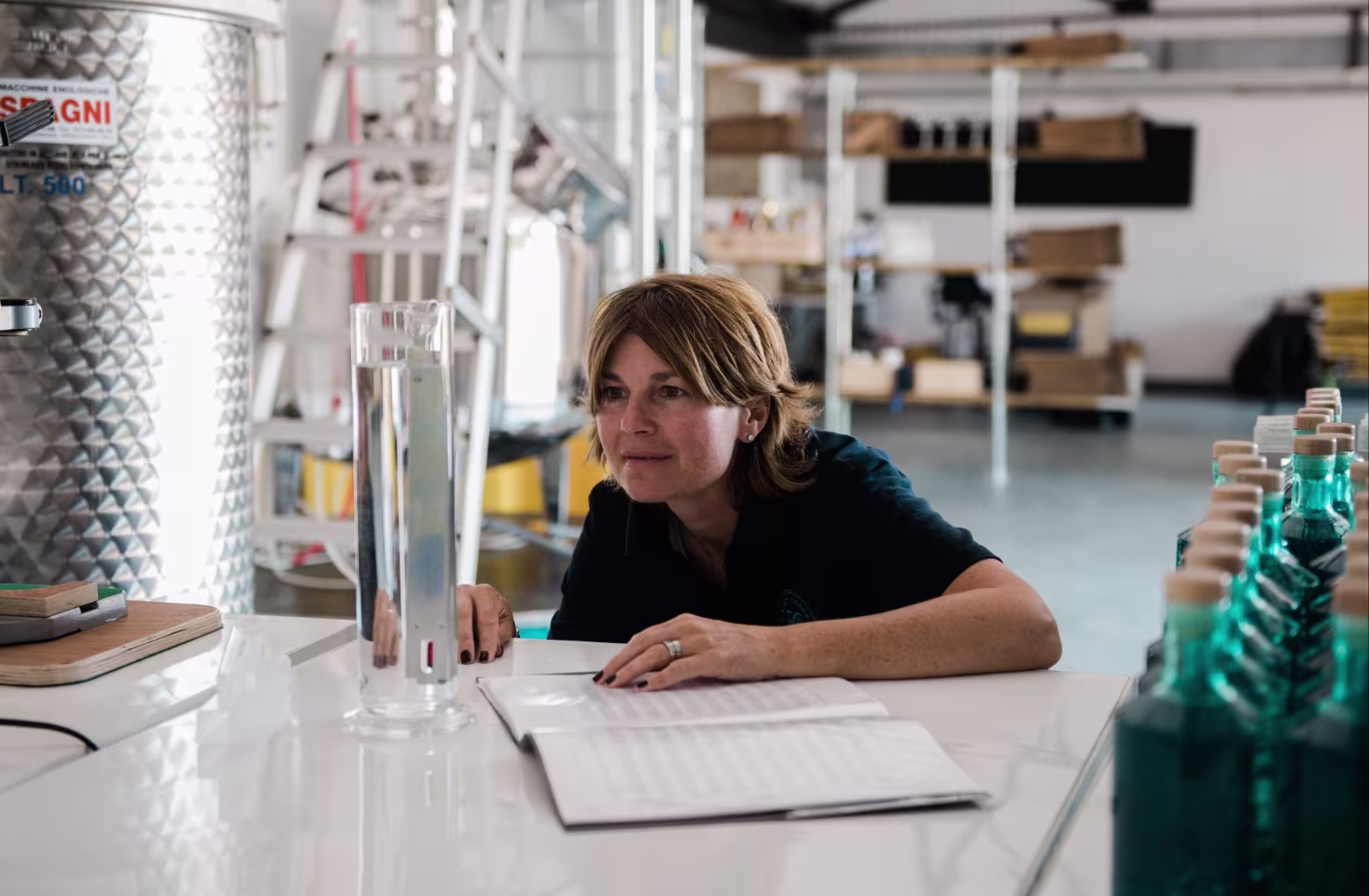 Woman closely examining a tall liquid measuring cylinder on a white table in an industrial setting.