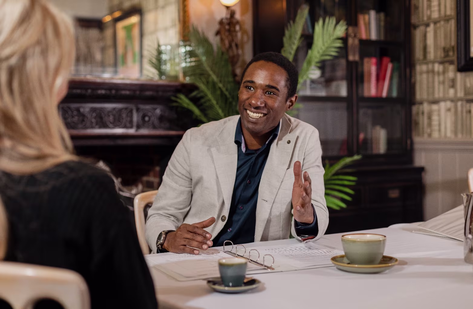 Man in beige jacket and blue shirt smiling and gesturing while talking to a woman at a table with coffee.