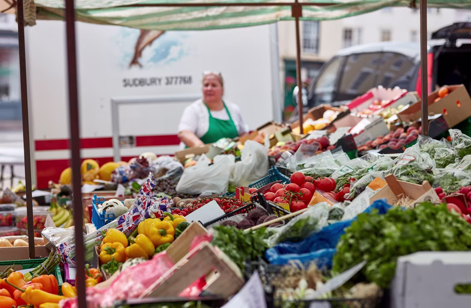 Woman in green apron stands behind market stall with fresh vegetables and fruits outdoors.