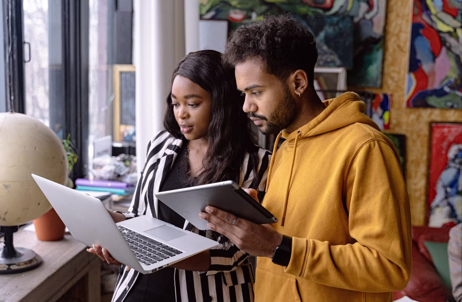 Two young adults looking at a laptop and a tablet in a room with paintings and a globe.