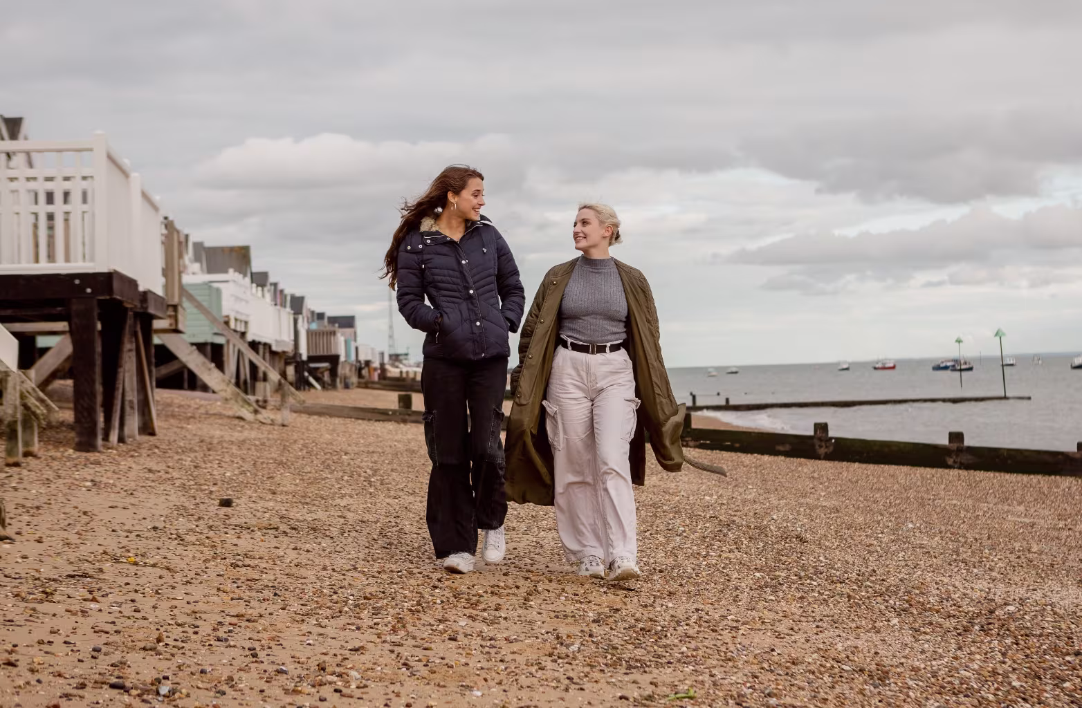 Two women walking and talking on a pebble beach near houses on stilts under cloudy sky.