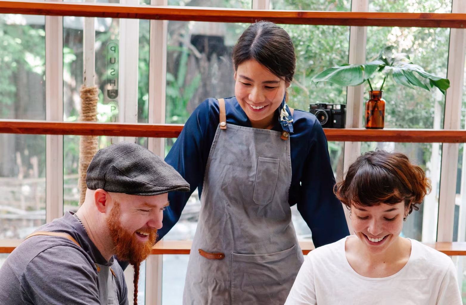 Three people smiling and chatting inside a bright room with large windows and plants.