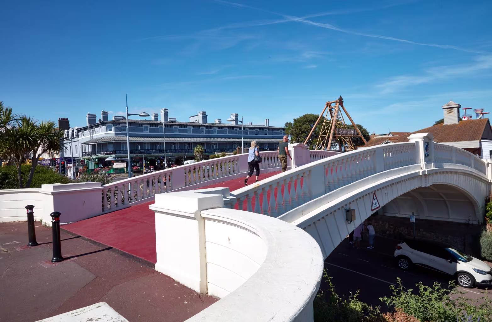 White pedestrian bridge with red walkway over a road, two people walking across under blue sky.