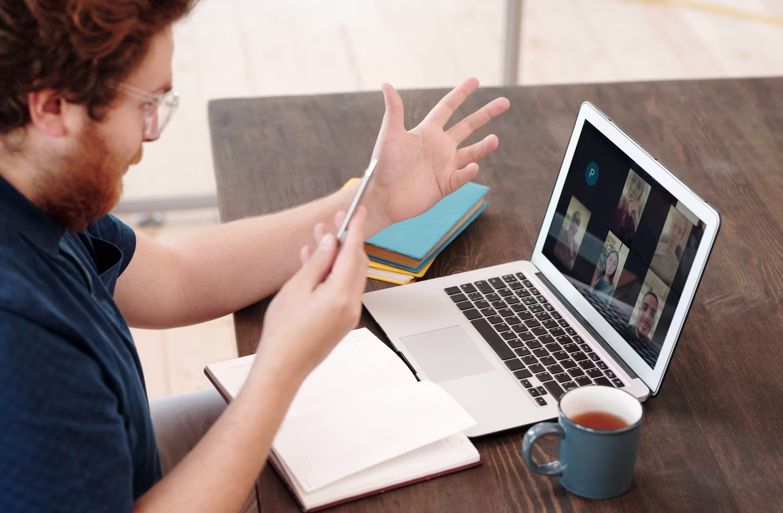Man with curly hair and glasses gestures during video call on laptop at wooden table with notebook and cup.