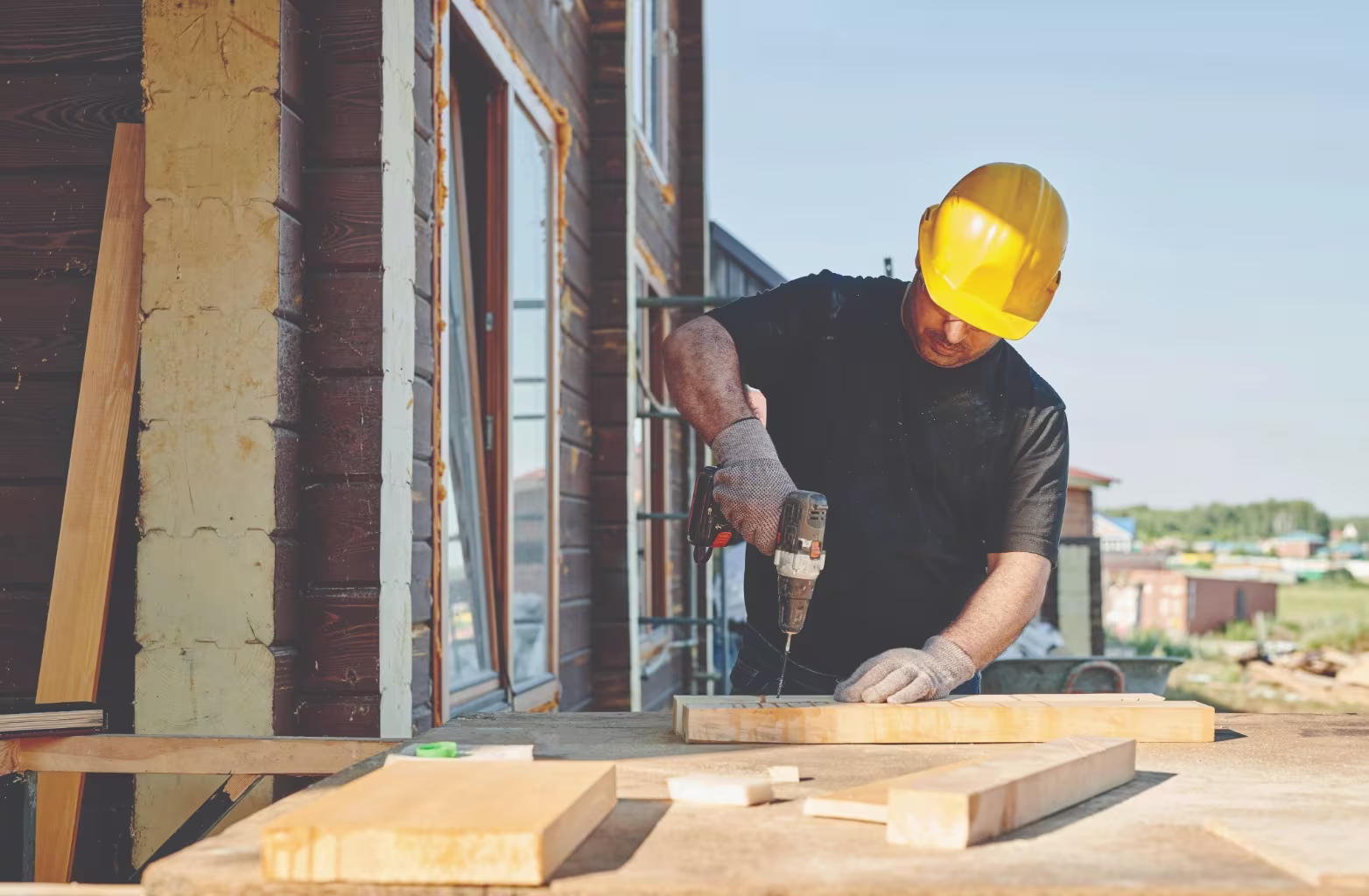 Construction worker wearing a yellow hard hat using a drill on a wooden plank outdoors.