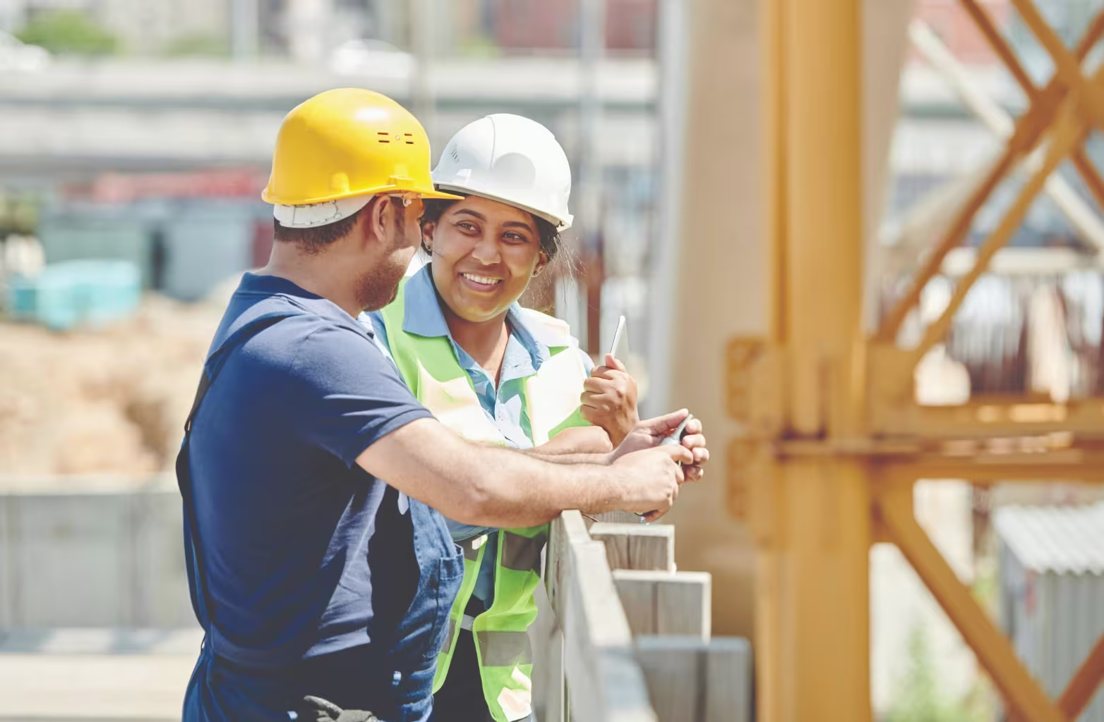 Two construction workers wearing helmets and safety gear smiling and talking at a construction site.