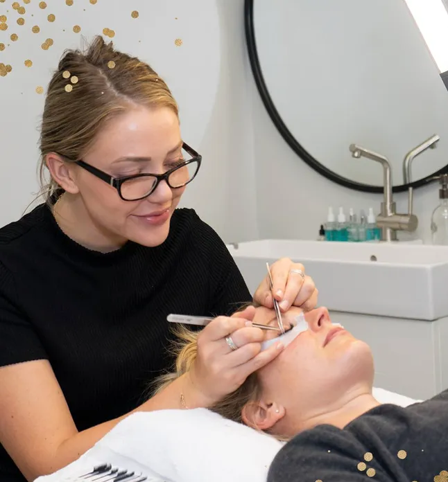 Esthetician wearing glasses applying eyelash extensions to a client lying on a treatment bed.