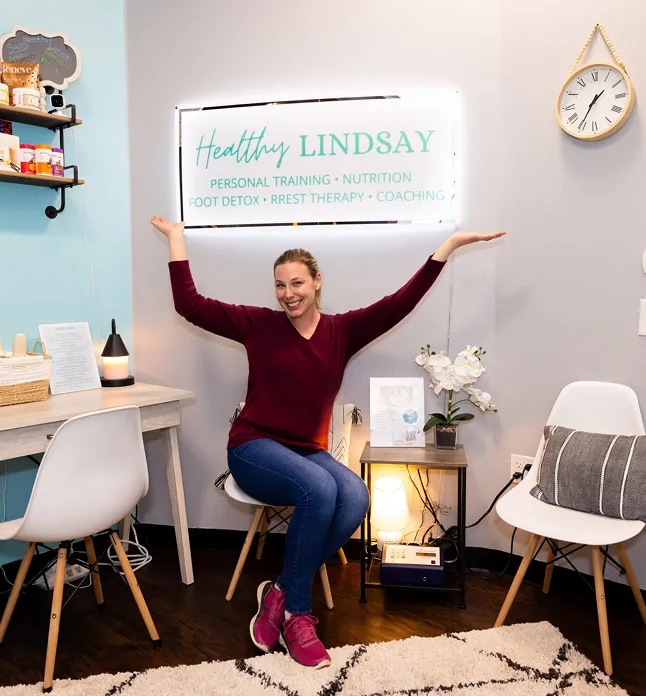 Woman in maroon sweater and jeans sitting on chair smiling, raising arms under Healthy Lindsay sign in wellness room.