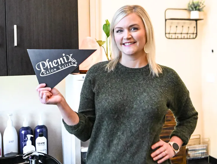 Smiling woman in green sweater holding a Phenix Salon Suites sign in a salon setting.