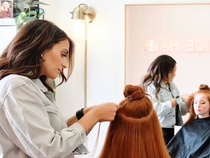 Hairdresser styling a client's long red hair in a salon with a mirror showing their reflections.
