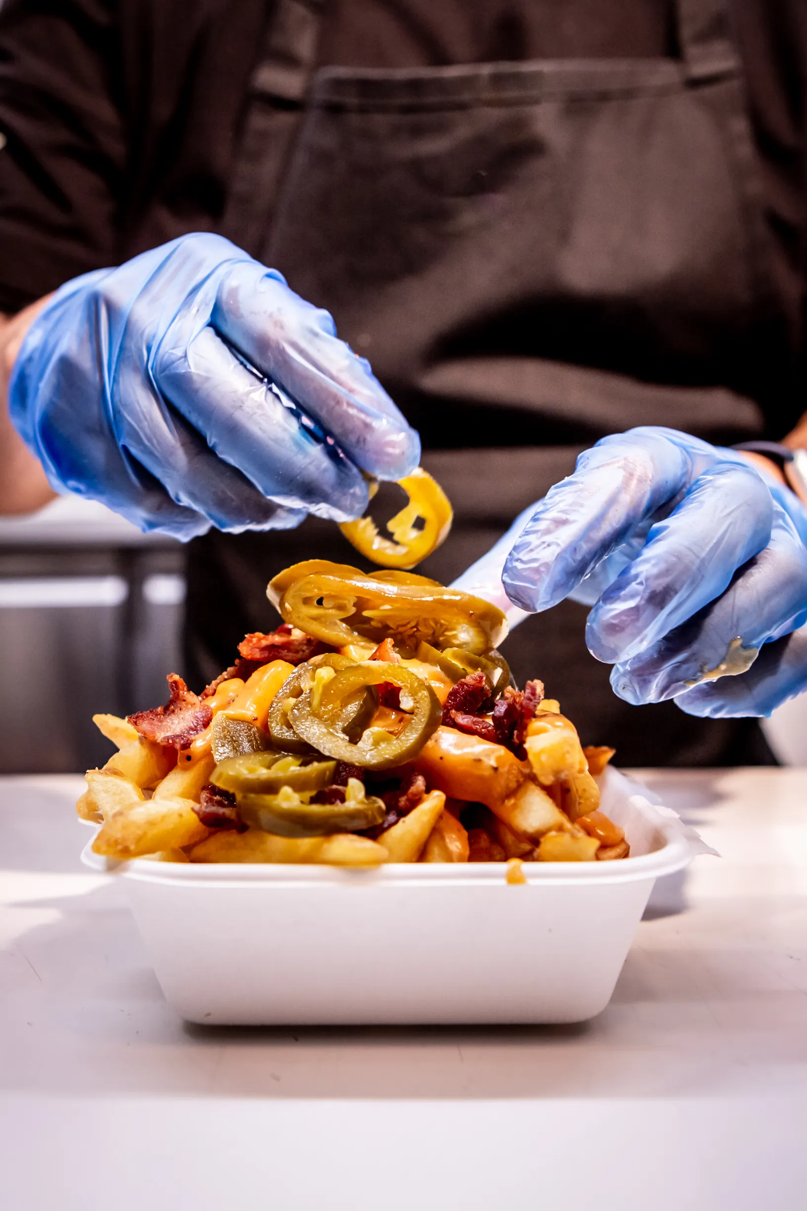 Hands with blue gloves adding sliced jalapeños on loaded fries with cheese and bacon in a white container.