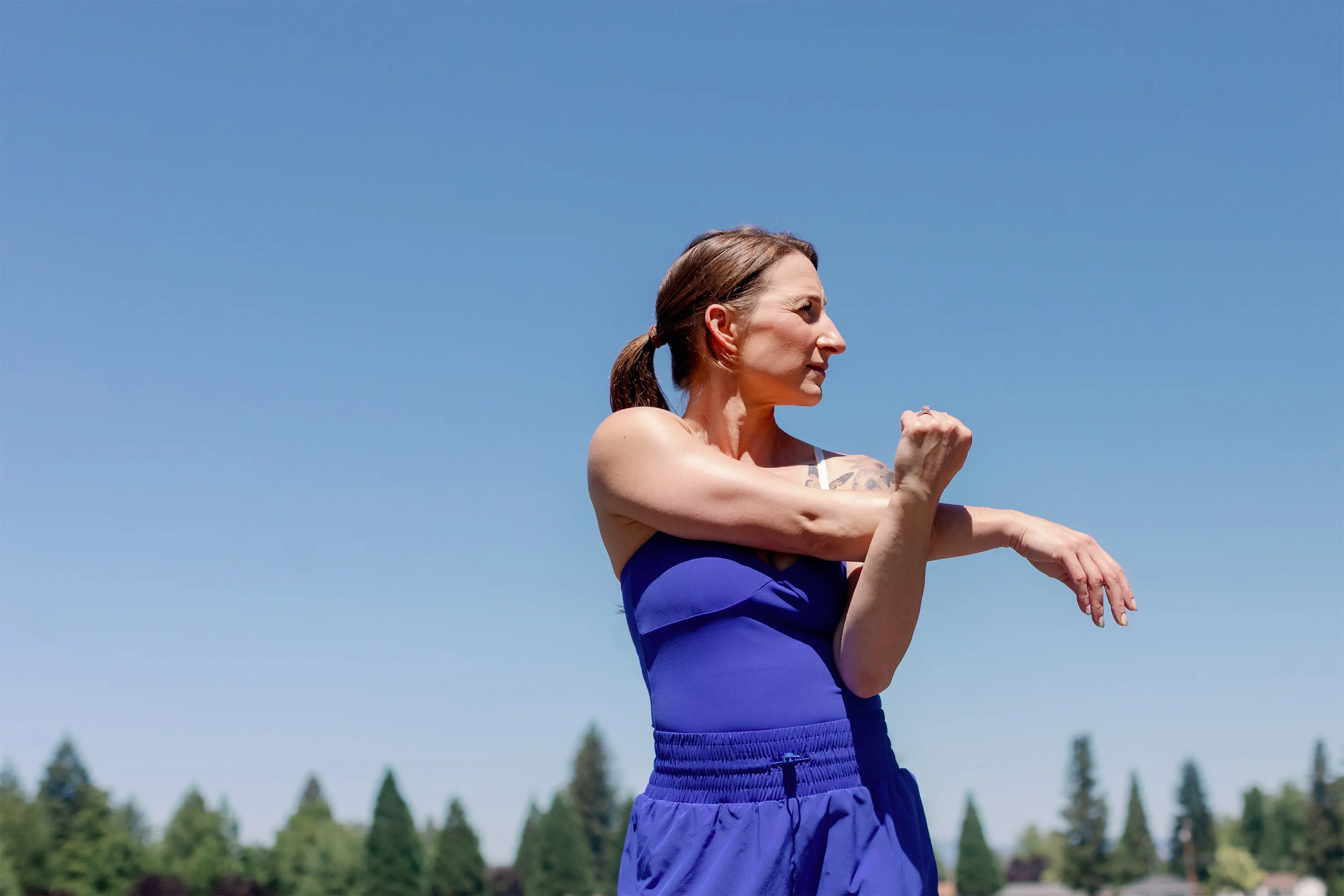 Woman in blue athletic wear stretching her arm with trees and clear sky in the background.