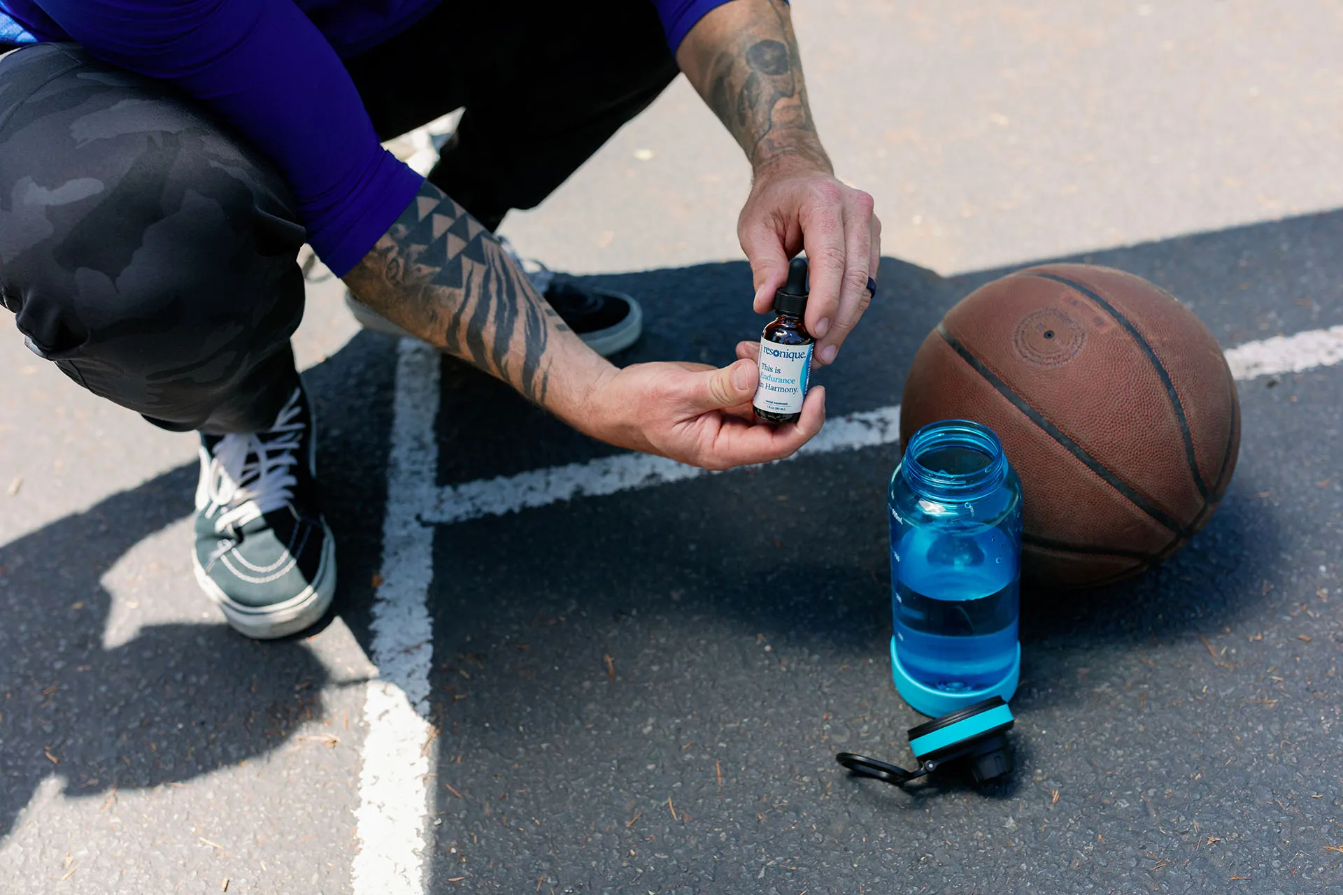 Tattooed person squatting holding a dropper bottle near a basketball and a blue water bottle on asphalt.