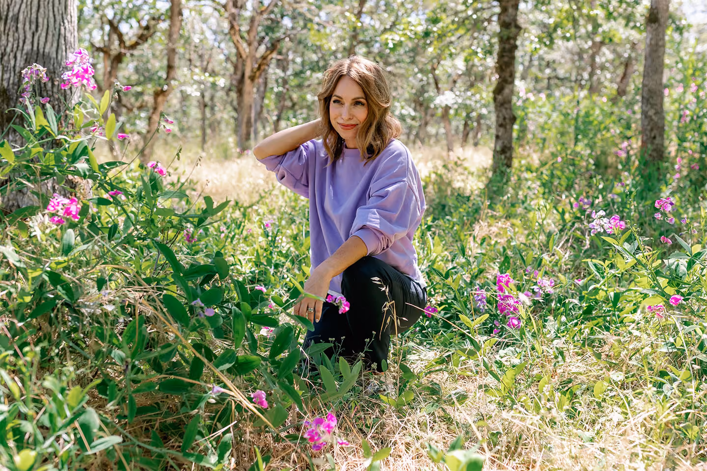 Woman in a purple sweater kneeling among green plants with pink flowers in a sunlit forest clearing.