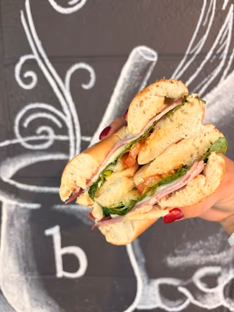 Hand holding a sandwich with ham, lettuce, tomato, and melted cheese against a dark wall with chalk art.