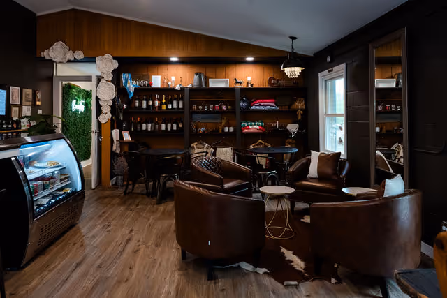 Interior cozy lounge area of bocaditos cafe with brown leather chairs, wooden shelves with bottles and pillows, and a glass display case of desserts.