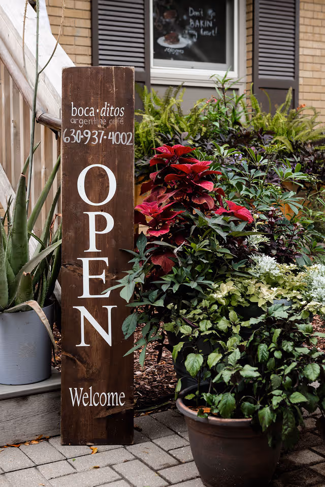 Wooden sign reading 'OPEN Welcome' with plants around, outside Bocaditos Argentine Cafe.