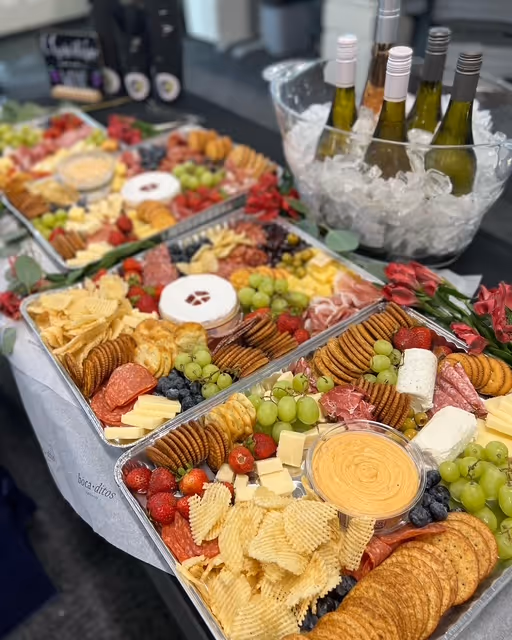 Three large trays of assorted cheeses, crackers, grapes, berries, and charcuterie with a bucket of white wine bottles on ice.
