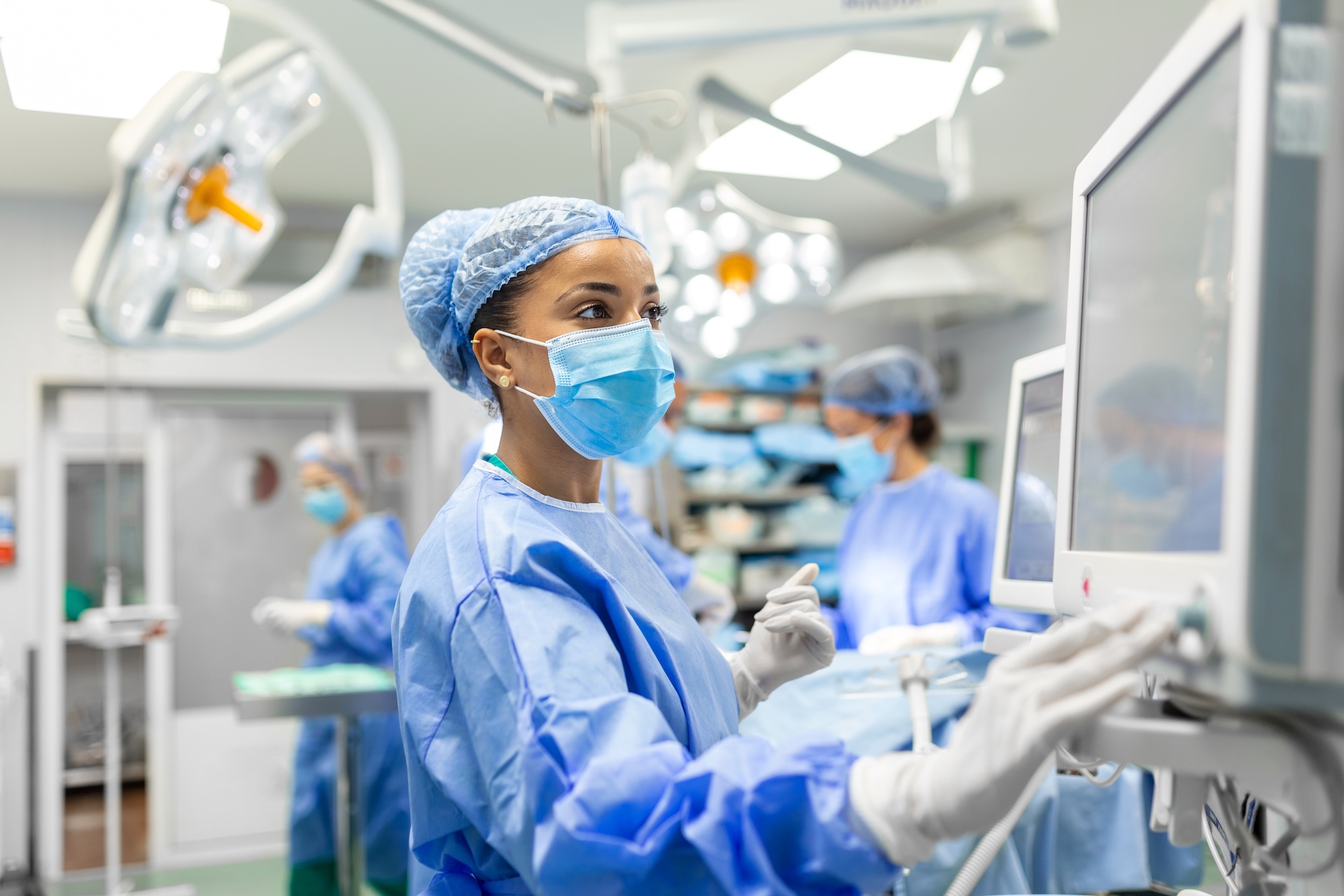 Female medical professional in surgical attire operating a monitor in a busy operating room.