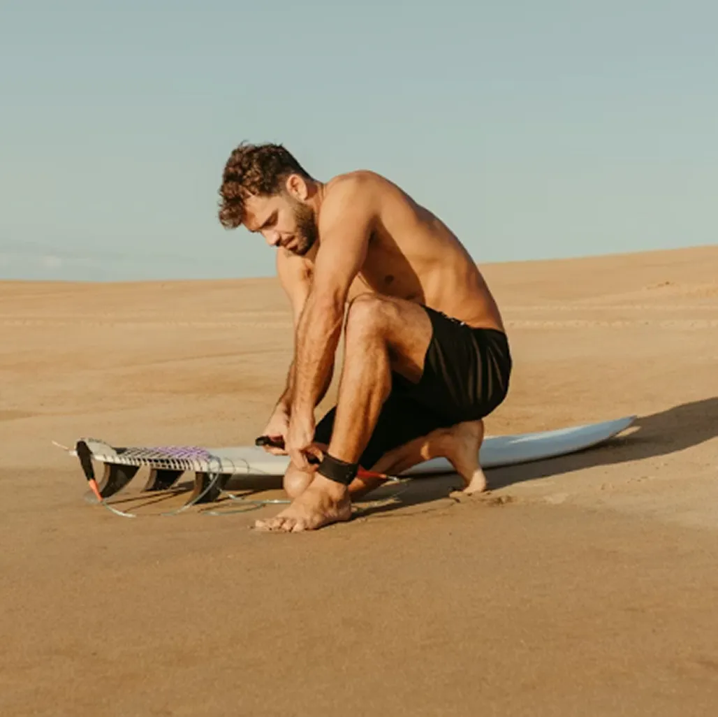 Shirtless man in black shorts fastening surfboard leash on sandy beach under clear sky.