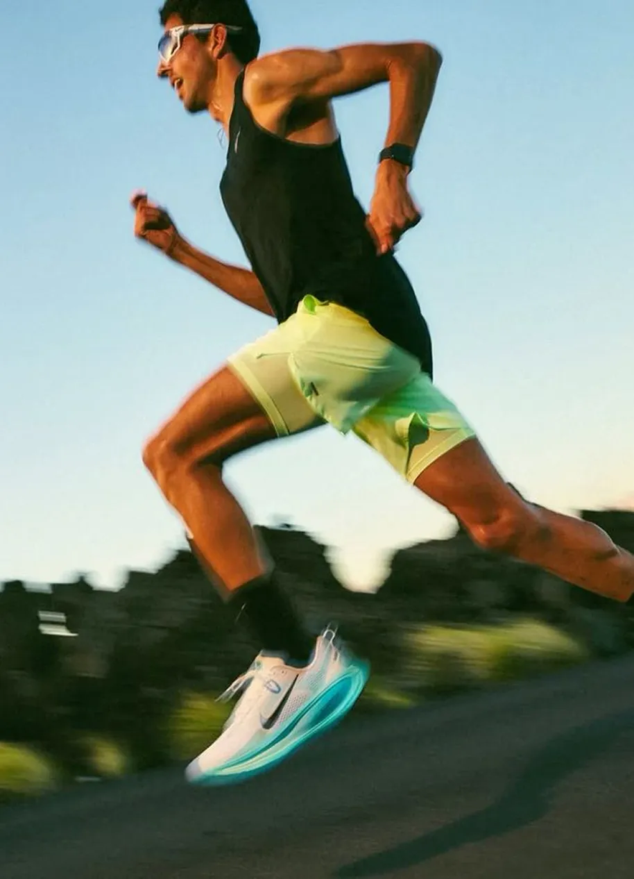 Man wearing sunglasses and neon shorts running outdoors in white Nike shoes with turquoise soles.