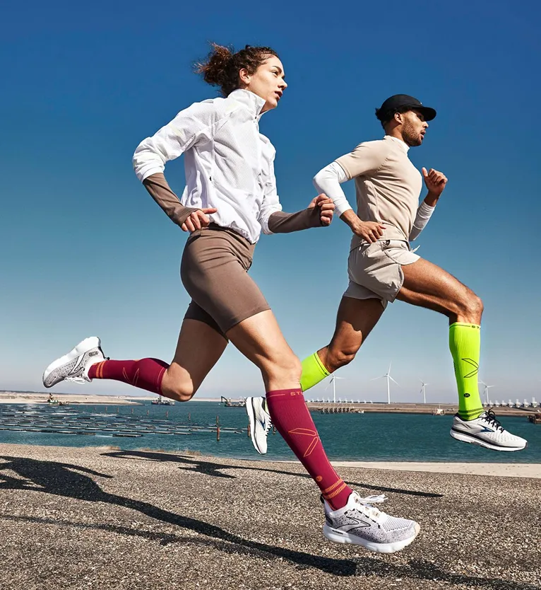 Two runners mid-stride with colorful compression socks running outdoors near water and wind turbines.