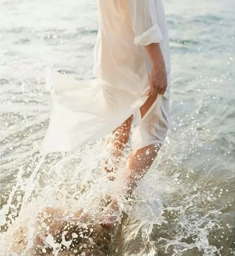 Person in white dress walking barefoot through splashing water at the edge of a body of water.