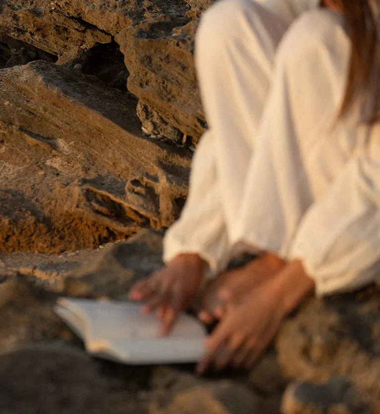 Person sitting on rocky ground with knees drawn up, holding an open book.