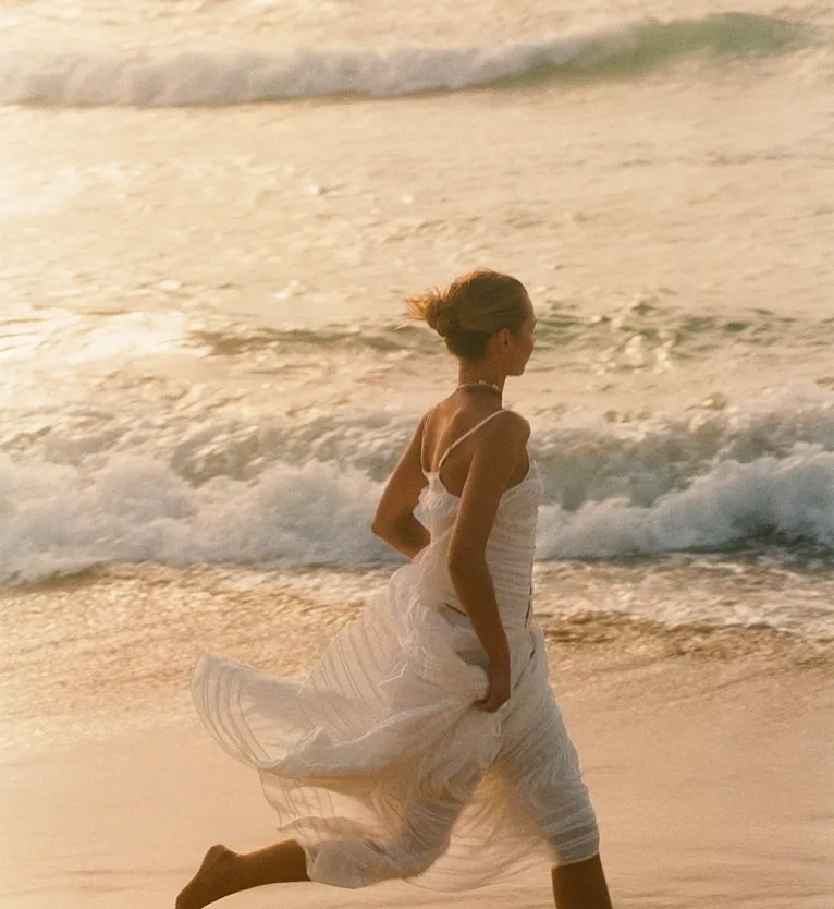 Woman in a white dress running barefoot along the beach near waves at sunset.