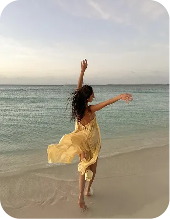 Woman in a flowing yellow dress dancing barefoot on a sandy beach near calm ocean waves.