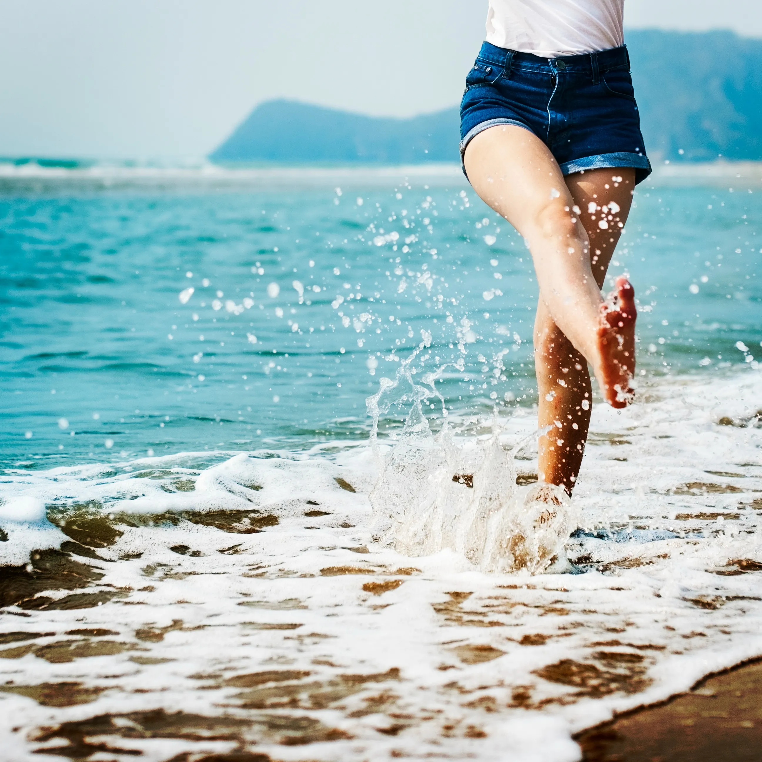 Person wearing denim shorts splashes water with one foot while standing in shallow ocean waves.