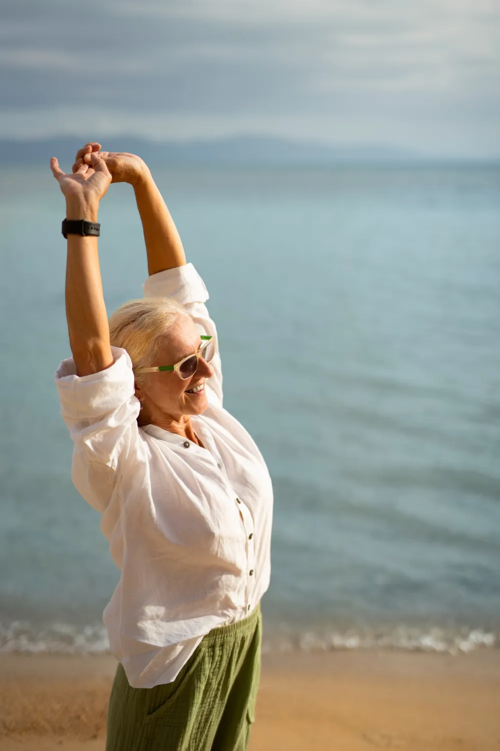 Smiling older woman wearing sunglasses stretching arms up at a beach with calm water background.